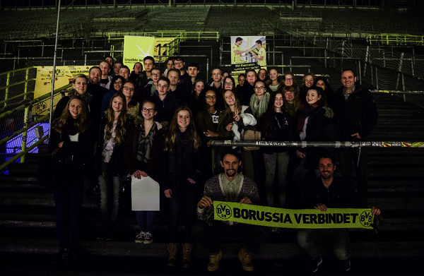 Ein Gruppenfoto der DGB-Jugend Dortmund auf der leeren Tribüne eines Fußballstadions. Zwei Menschen, die in der ersten Reihe hocken haltenen einen BVB-Schal mit der Aufschrift "Borussia verbindet" hoch.