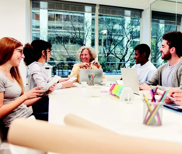 Ein Foto einer Seminarsituation mit Erwachsenen. Fünf Menschen sitzen an einem Tisch, auf dem Laptops und Schreibwaren stehen. Sie lächeln sich gegenseitig an.