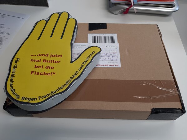 Ein Foto von einem Paket, das auf einem Schribtisch liegt. Auf dem Paket liegt ein Stapel großer ausgeschnittener Papierhände. Bei den Händen handelt es sich um das Logo der Gelben Hand. Mittig auf der Handfläche steht in roter Schrift: "... und jetzt mal Butter bei die Fische!"