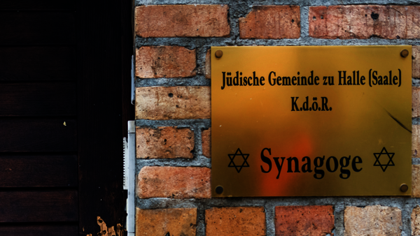 Ein Foto von einer goldenen Tafel, die an einer Backsteinwand hängt mit der Aufschrift "Jüdische Gemeinde zu Halle (Saale) K.d.ö.R Synagoge". Links daneben sind dunkelbraune Blendläden zu sehen.