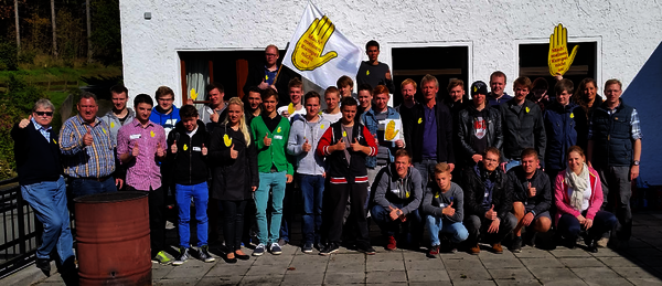Ein Gruppenfoto von Auszubildenden der Rheinbahn AG draußen im Sonnenschein vor einem Haus. Einige haben Gelbe Hand Anstecker oder eine Fahne der Gelben Hand. Viele heben einen Daumen.