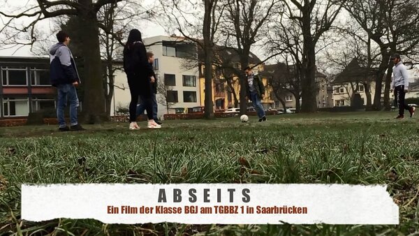 Ein Foto aus der Froschperspektive von jungen Menschen, die auf Rasen Fußballspielen. Im Hintergrund sind Häuser zu sehen.