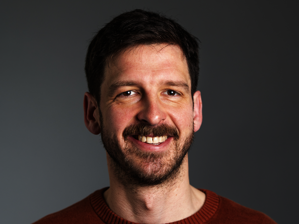 Ein Proträtfoto von Roland Maas. Roland Maas trägt einen blau und orangenen Pullover und lächelt breit in die Kamera, sodass seine Zähne zu sehen sind.